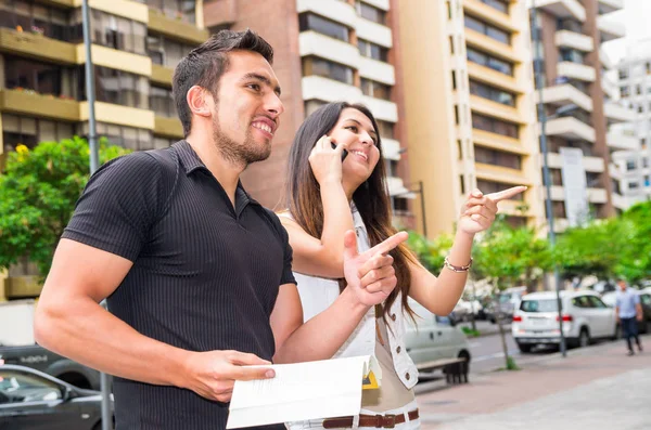 Charming young couple standing outside in urban environment, holding open book and woman talking on mobile interacting, urban tourist concept — Stock Photo, Image