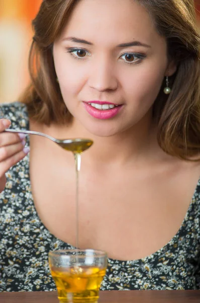 Junge Frau vor der Kamera hält Löffel mit Honig, der in ein kleines Glas auf dem Tisch tropft — Stockfoto