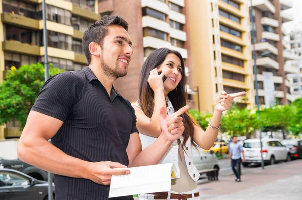 Charming young couple standing outside in urban environment, holding open book and woman talking on mobile interacting, urban tourist concept — Stock Photo, Image