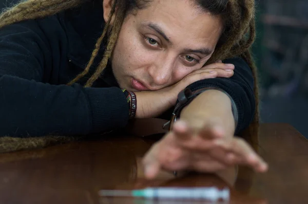 Man lying over table with dopey facial expression, reaching for syringe across desk, drug addiction concept — Stock Photo, Image