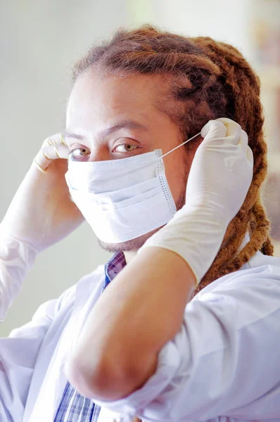 Médico joven con largos bloqueos de terror posando para la cámara, ajuste de la máscara facial que cubre la boca, clínica en el fondo, concepto médico — Foto de Stock