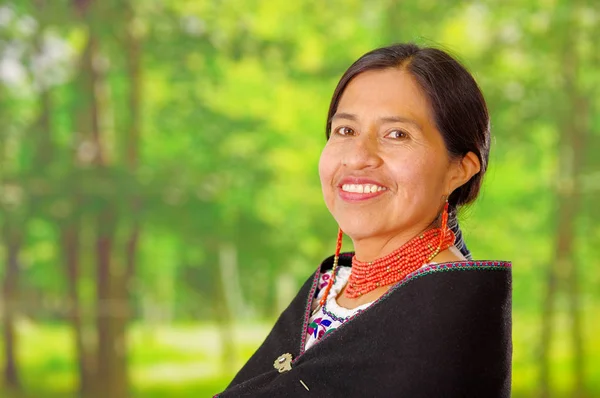 Closeup beautiful hispanic woman wearing traditional andean white blouse with colorful decoration around neck, black poncho, matching red necklace and ear, posing happily for camera, garden background