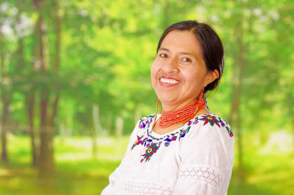 Closeup beautiful hispanic woman wearing traditional andean white blouse with colorful decoration around neck, matching red necklace and ear, posing happily for camera, garden background