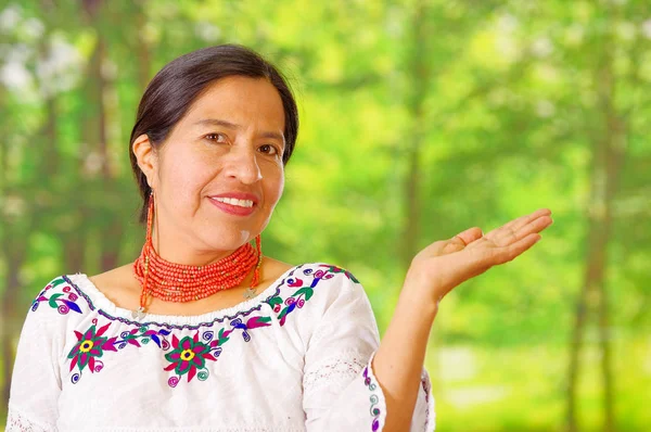 Feche a bela mulher hispânica vestindo blusa branca andina tradicional com decoração colorida ao redor do pescoço, combinando colar vermelho e orelha, posando feliz para a câmera segurando palma da mão, jardim — Fotografia de Stock