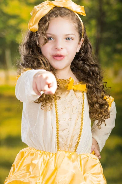 Carino bambina indossa bel vestito giallo con fascia abbinata testa, in posa per la fotocamera, sfondo verde foresta — Foto Stock