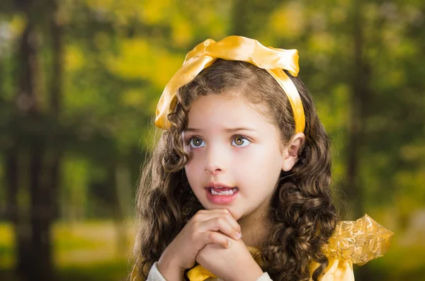 Headshot cute little girl wearing beautiful yellow dress with matching head band, posing for camera, green forest background — Stock Photo, Image