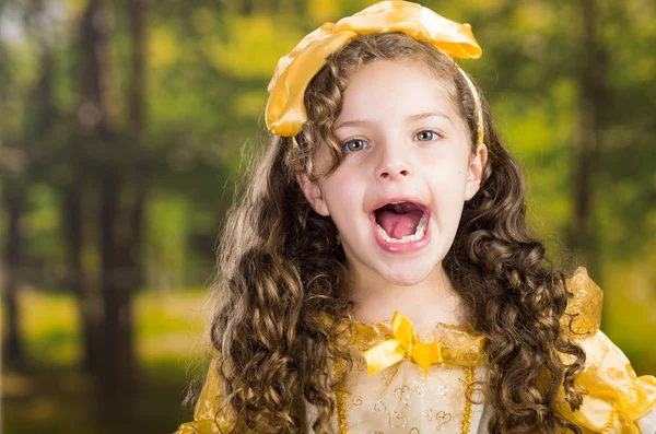 Headshot carina bambina indossa bel vestito giallo con fascia abbinata testa, in posa per la fotocamera, sfondo verde foresta — Foto Stock