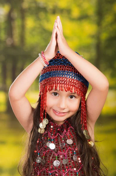 Petite fille mignonne portant une belle robe rouge avec un chapeau de perles assorti, posant pour appareil photo, fond de forêt verte — Photo