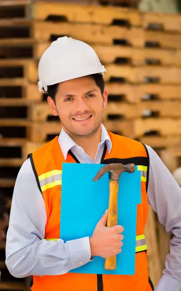 Sonriente joven ingeniero sosteniendo una carpeta y martillo en su mano en el sitio de construcción —  Fotos de Stock