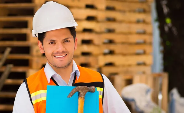 Sorrindo jovem engenheiro segurando uma pasta e martelo em sua mão no canteiro de obras — Fotografia de Stock