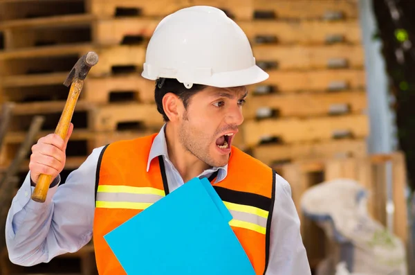 Schöner junger Ingenieur mit Aktenordner und Hammer in der Hand auf der verrückten Baustelle — Stockfoto