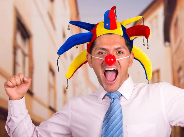 Feliz joven hombre de negocios guapo con una nariz de payaso rojo y cabaña de arlequín colorido en su cabeza — Foto de Stock