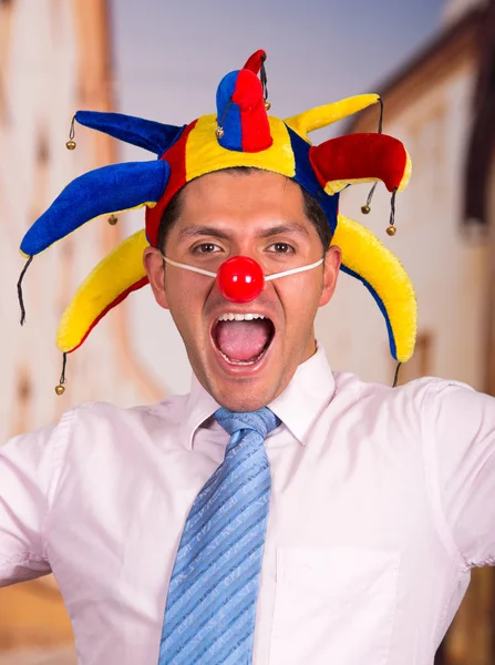 Glad stilig ung affärsman med röd clown näsa och färgglada harlequin hut i huvudet — Stockfoto