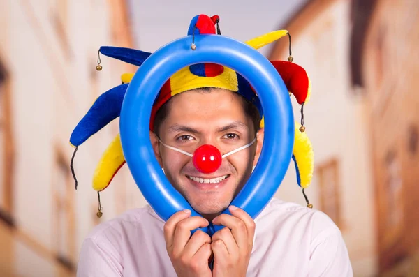 Giovane uomo d'affari allegro con un naso da clown rosso e una capanna colorata arlecchino nella sua testa che gioca con un palloncino blu intorno alla testa — Foto Stock