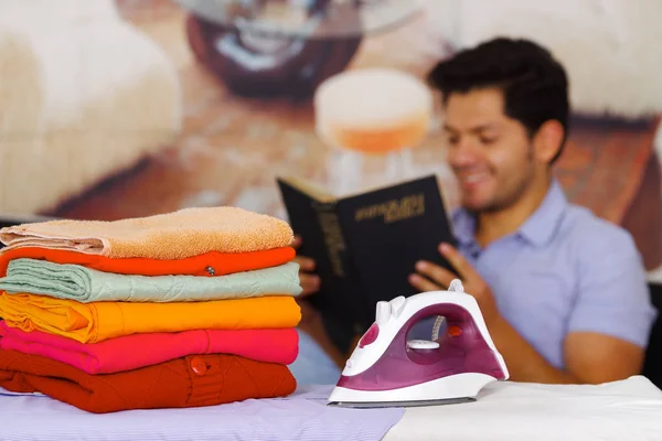 Un mucchio di vestiti accanto al ferro da stiro con splendido uomo sorridente dietro mentre sta leggendo un libro — Foto Stock