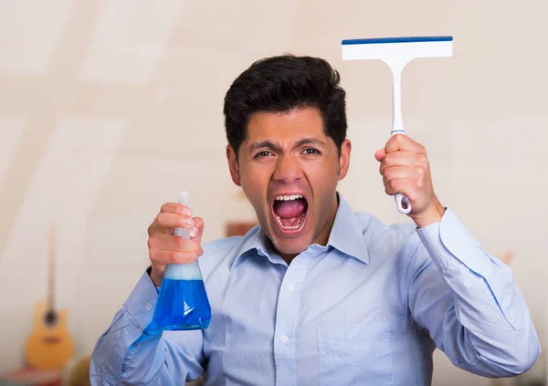 Bonito jovem caucasiano masculino segurando uma garrafa de spray de limpeza em uma mão e um limpador de janelas em sua outra mão enquanto ele está gritando — Fotografia de Stock