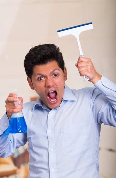 Handsome young caucasian male holding a cleaning spray bottle in one hand and a window cleaner in his other hand while he is screaming — Stock Photo, Image