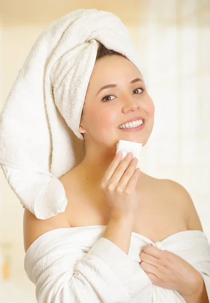 Bela jovem sorridente com uma toalha branca cobrindo sua cabeça está limpando seu queixo com um pequeno pedaço de papel no banheiro — Fotografia de Stock
