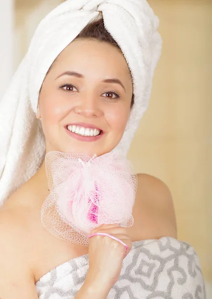 Bela jovem sorridente com toalha branca cobrindo sua cabeça colocar uma esponja de banho rosa sob sua bochecha — Fotografia de Stock