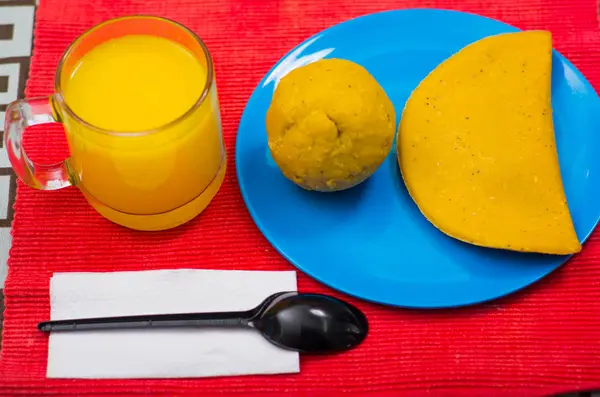 Um café da manhã energético e completo contém suco de laranja, empanada e bolon servido em um prato azul, com uma colher preta sobre um guardanapo. Conceito tradicional de comida andina — Fotografia de Stock