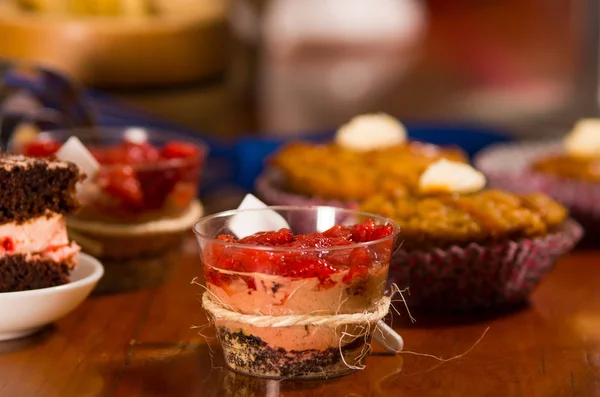 Deliziosa mousse ricoperta di topping alla fragola, presentata all'interno di piccoli bicchieri con cucchiai attaccati, pezzi da torta e fondo muffin, concetto di pasticceria — Foto Stock