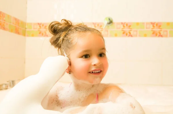 Little beautiful girl touching her ear with her hand inside of a bathtub with water and foam — Stock Photo, Image