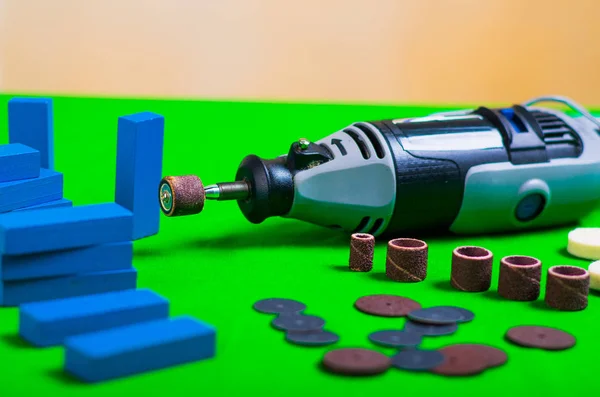 Un trapano grigio con alcuni accessori di perforazione con un piccolo pezzi di legno blu su uno sfondo verde — Foto Stock