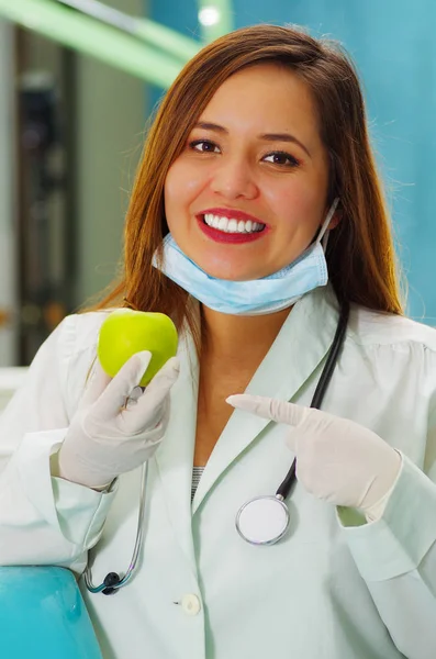 Beautiful woman patient with a stethoscope around her neck is holding a delicious apple in a dentists office — Stock Photo, Image