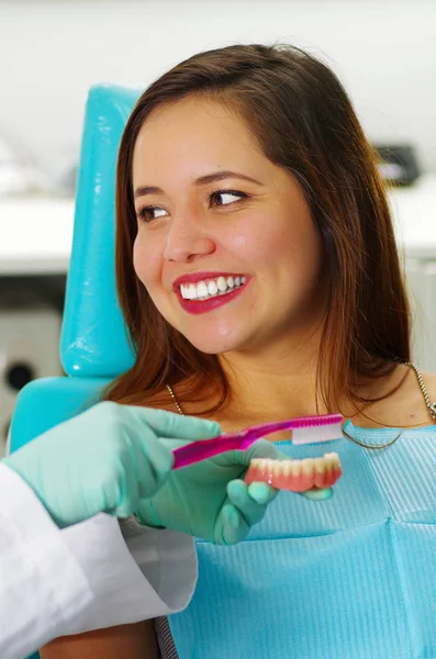 Mulher bonita paciente sorrindo enquanto o médico está escovando uma placa dental falsa em um consultório de dentistas — Fotografia de Stock