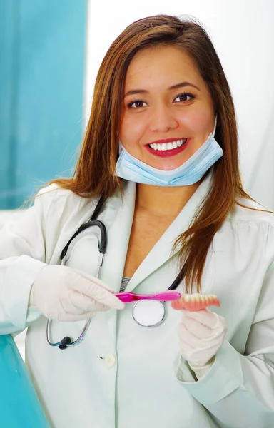 Красивая женщина-врач позирует перед камерой, используя хирургическую маску на подбородке, пока она чистит поддельную зубную табличку — стоковое фото