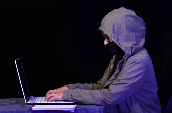 Retrato de um hacker menina bonita e inteligente com laptop no fundo escuro usando óculos e cobrindo seu pescoço, boca e nariz com sua camisola gola alta e cobrindo a cabeça com uma jaqueta — Fotografia de Stock