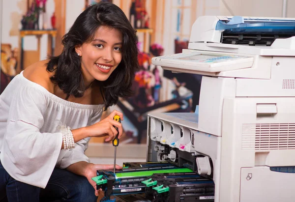 Mooie vrouw dragen een witte bloes, gehurkt en tot vaststelling van een fotokopieerapparaat en glimlachen tijdens het onderhoud met behulp van een schroevendraaier — Stockfoto