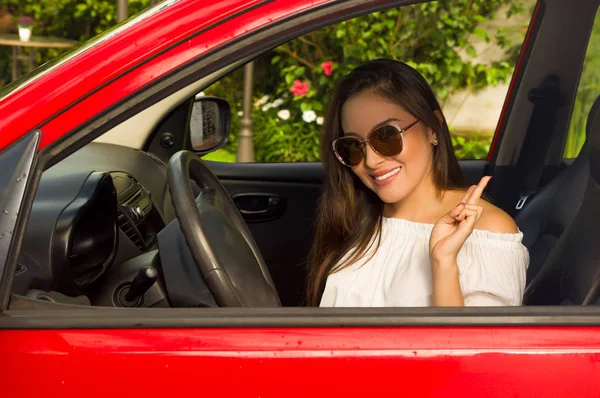 Ładna dziewczyna sobie okulary i robi sygnału zwycięstwo z jej strony wewnątrz jej czerwony samochód — Zdjęcie stockowe