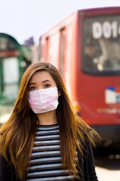 Mladá žena s ochrannou maskou na ulici ve městě s znečištění ovzduší, rozmazané pozadí veřejné dopravy — Stock fotografie