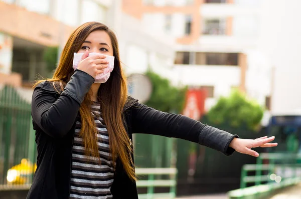 Mujer joven sosteniendo su máscara protectora con la mano en la calle de la ciudad con la contaminación del aire, pidiendo un taxi, fondo de la ciudad — Foto de Stock