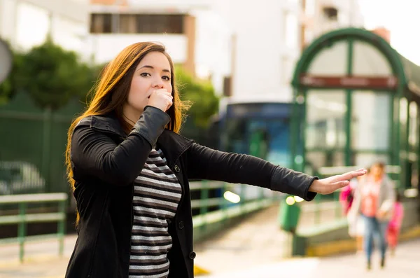 Mujer joven con máscara protectora cubriéndose la boca con una mano y pidiendo un taxi con su otra mano en la calle de la ciudad con contaminación del aire, fondo de la ciudad —  Fotos de Stock