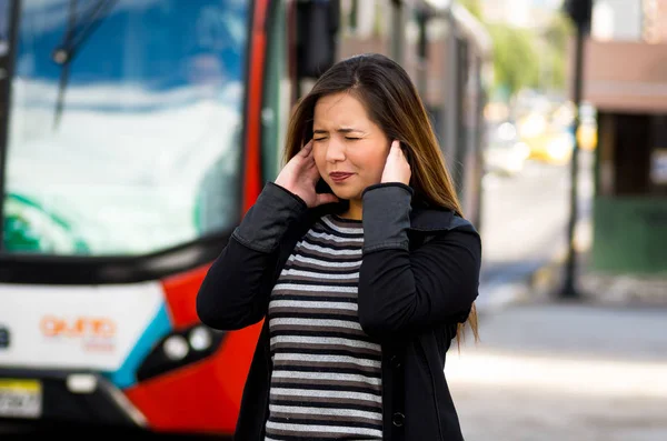 Primo piano di una bella giovane donna felice che si copre le orecchie con entrambe le mani per strada in città con inquinamento acustico con un autobus sfocato dietro, sfondo della città — Foto Stock