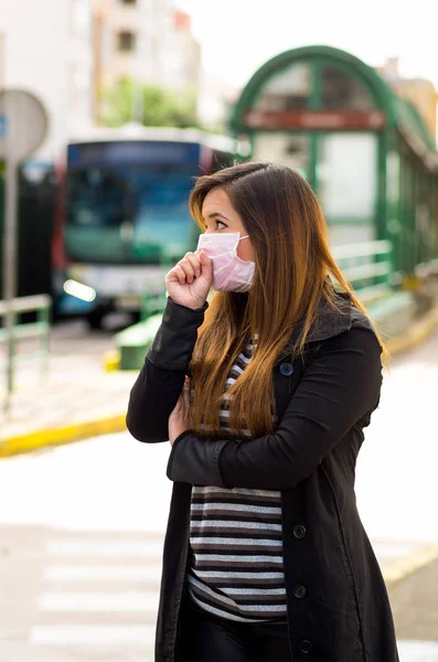 Mujer joven con máscara protectora tosiendo en la calle de la ciudad con contaminación del aire, fondo borroso del transporte público — Foto de Stock