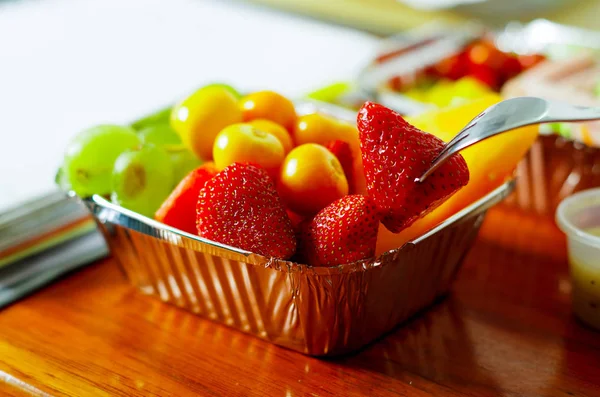 Healthy man using a fork to eat in the office a delicious fresh fruit salad on aluminium box on wooden table — Stock Photo, Image