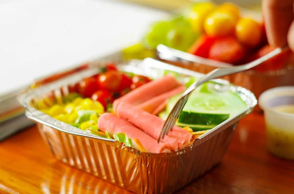 Homem saudável usando um garfo para comer no escritório uma deliciosa salada fresca de estilo mediterrâneo com presunto na caixa de alumínio na mesa de madeira — Fotografia de Stock