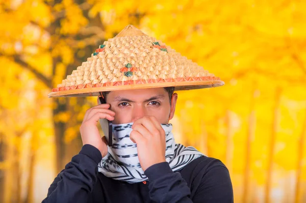 英俊的西班牙裔年轻商业头脑的人穿亚洲圆锥形的帽子，遮住他的脸，用一条围巾以及在秋天背景使用他的手机的肖像 — 图库照片