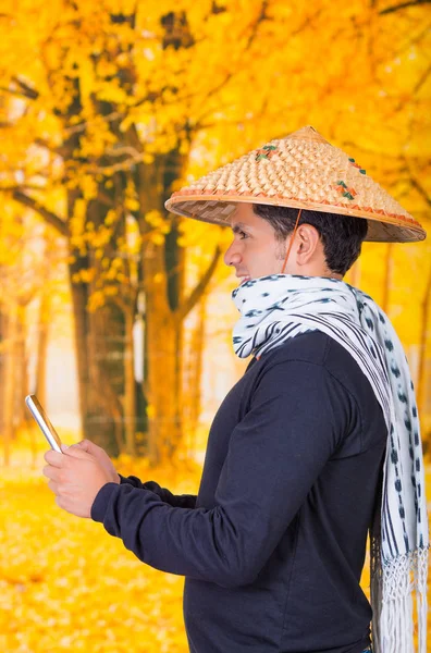 Porträtt av en stilig spansktalande unga företag kille klädd i en asiatisk konisk hatt och en scarf runt halsen håller sin Ipad med båda händerna, i hösten bakgrund — Stockfoto