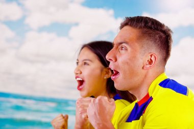 Resmi maraton futbol gömlek ayakta dış yüzey fotoğraf makinesi, büyük bir coşku, mavi gökyüzü ve bulutlar arka plan ile çok meşgul vücut dili izlerken oyun giyen genç Ekvadorlu Çift