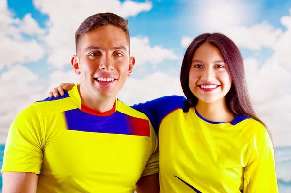Giovane coppia ecuadoriana indossa ufficiale maratona di calcio camicia in piedi di fronte alla fotocamera, molto impegnato linguaggio del corpo a guardare il gioco con grande entusiasmo, cielo blu e nuvole di sfondo — Foto Stock
