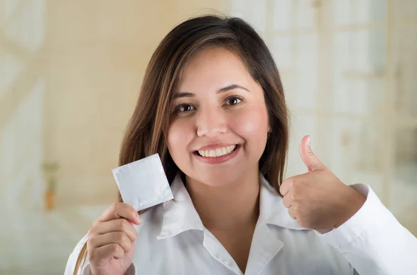 Young beautiful doctor holding a condom package and doing with her hand a thumb up, safe sex concept. Protection against AIDS and birth control — Stock Photo, Image
