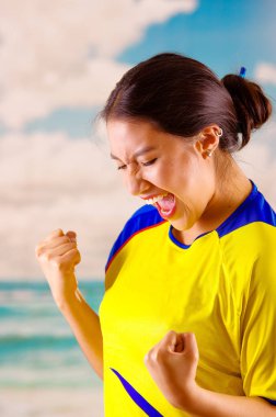 Resmi maraton futbol gömlek ayakta dış yüzey fotoğraf makinesi, büyük bir coşku, mavi gökyüzü ve bulutlar arka plan ile çok meşgul vücut dili izlerken oyun giyen genç Ekvadorlu kadın