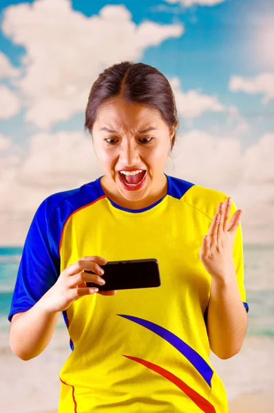 Tânăra femeie ecuadoriană purtând tricou oficial de fotbal maraton în picioare cu care se confruntă camera, foarte angajat limbajul corpului vizionarea joc cu mare entuziasm, cer albastru și nori fundal — Fotografie, imagine de stoc