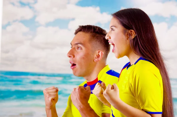 Jeune couple d'écuadoriens portant une chemise officielle de football Marathon debout face à la caméra, un langage corporel très engagé regardant le jeu avec beaucoup d'enthousiasme, un ciel bleu et un fond nuageux — Photo