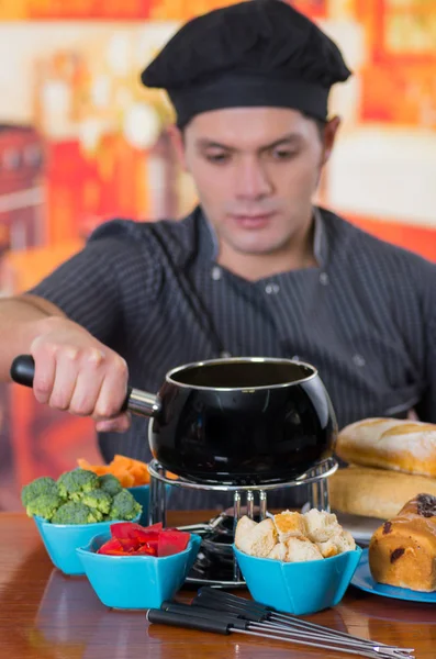 Unga cheff förbereder en schweizisk fondue gourmetmiddag med ostar och en uppvärmd gryta med ostfondue och vissa grönsaker som broccoli, morot och paprika och brödbitar insidan skålar — Stockfoto