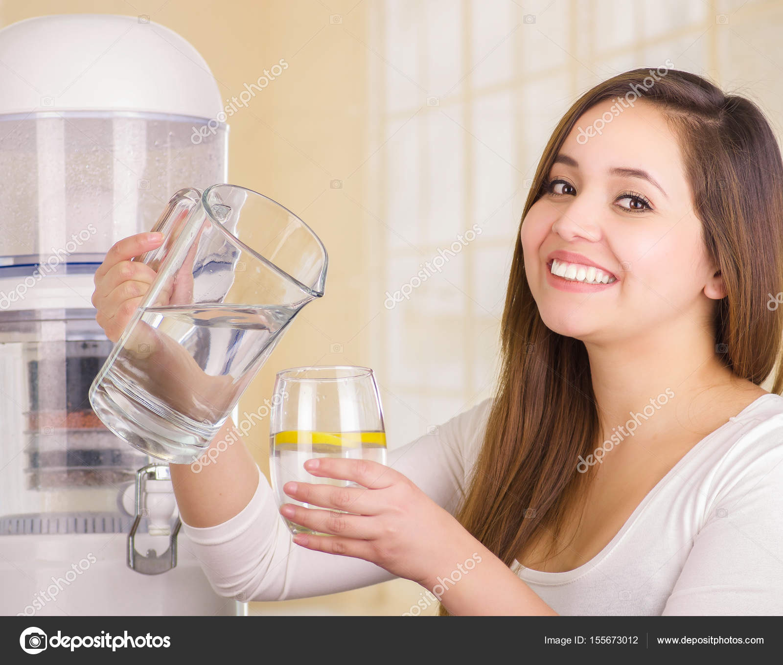 Beautiful smiling woman holding a glass of water in one hand and a pitcher  of water in her other hand, with a filter system of water purifier on a  kitchen background Stock